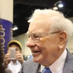 Warren Buffett smiling with a person taking his picture with a phone in the background.