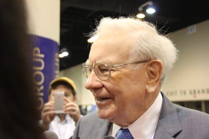 Warren Buffett smiling with a person taking his picture with a phone in the background.