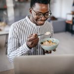 A person smiles while eating breakfast cereal with fruit in front of a laptop computer.